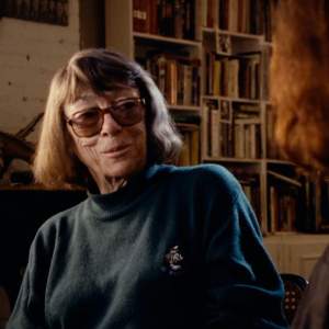 Joan Mitchell sits in a darkened room lined with bookshelves, facing an unseen interviewer. Mitchell has short brown hair, tinted glasses and light skin tone.