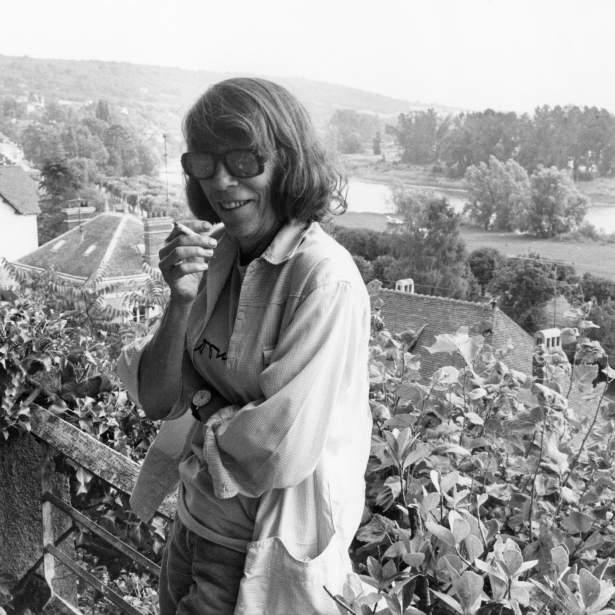In a black and white photo, artist Joan Mitchell smiles wearing dark glasses in front a sweeping landscape view.