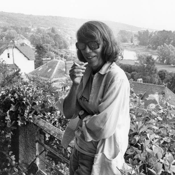 Joan Mitchell smiles and holds a cigarette, leaning against a railing, overlooking a lush landscape with trees and the river Seine. She is a white woman in her mid-50s, wearing sunglasses and a loose dress shirt, and has a shoulder length bob.