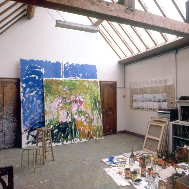 View of an art studio with beamed ceilings, large abstract paintings and art materials laid out on the floor.