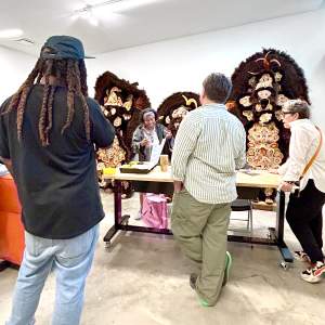 In front of a studio wall with 3 elaborate feathered and beaded costumes, 3 people visit Denice who stands at center,at center, a Black woman with short black hair.