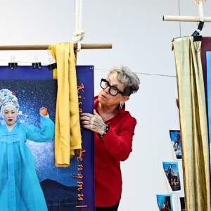 Jerri Allyn adjusts satin fabric on a handing figurative photomontage. She is a white woman with short blond hair, black frame glasses, red shirt, and skeletal tattoos on her hand.