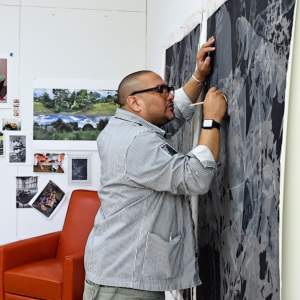 David Antonio Cruz draws with a white pencil on a large greyscale painting on paper tacked to the wall. Behind him, photographs of landscapes and artworks are on the wall. he is a Latino man with shaved head, and wears black frame glasses and a work jacket.
