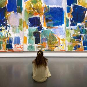 A woman with long curly hair sits on a gallery floor in front of a large multi-panel abstract painting, covered with rectilinear fields of blues and greens, punctuated by accents of orange and yellow.