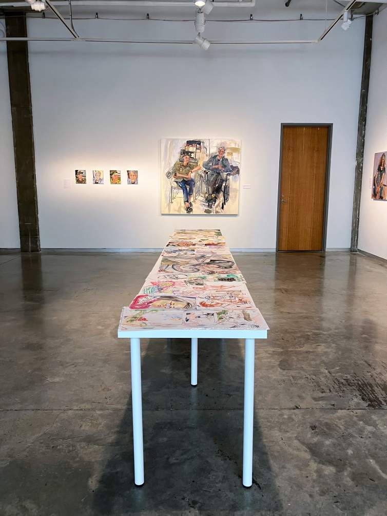In an art gallery with white walls and a gray concrete floor, figurative paintings in a gestural style hang on the walls. In the center of the room, a long table holds various sketches.