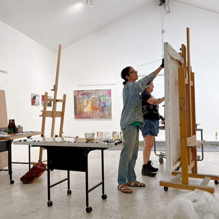 Artists Mala Iqbal and Angela Dufresne stand in front of a large canvas on an easel in an artist’s studio, painting together.