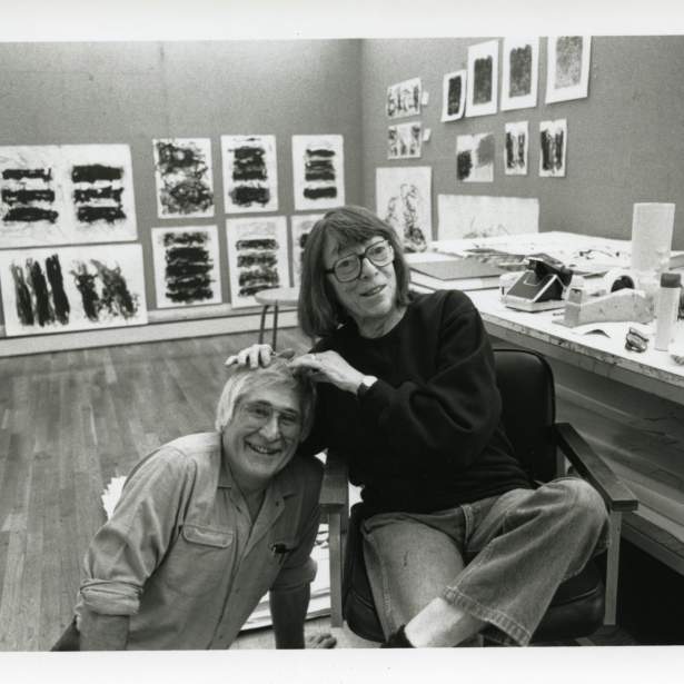 In a studio lined with proofs of abstract prints on the wall, Joan Mitchell rests her hands on Ken Tyler's head, who smiles and crouches on the floor next to her chair.